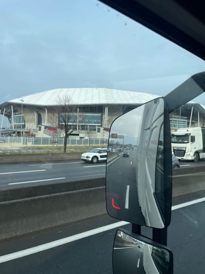 The image depicts an exterior scene from the perspective of a vehicle, likely a truck, showcasing a multi-lane highway and the Groupama Stadium in Décines-Charpieu, France, under an overcast sky.
In the background, the Groupama Stadium stands prominently, characterized by its distinctive white, ridged roof and a multi-tiered, grey architectural facade. The stadium's name, "Groupama Stadium," is partially visible on its exterior. Several leafless trees are situated between the stadium grounds and the highway, separated by a fence.
The midground features a dark asphalt highway with visible white lane markings. A concrete barrier separates the lanes of travel. Vehicles are present on the highway, including a white SUV (consistent with a Citroën C4 Cactus) in the lane adjacent to the barrier, traveling in the same direction as the viewpoint vehicle.
The foreground is dominated by the interior framing of the viewpoint vehicle's window at the top and right. A large side mirror, characteristic of a commercial truck, occupies the mid-right portion of the image. This mirror reflects the highway behind the viewpoint vehicle, showing additional cars and a white DAF semi-truck. A smaller, secondary mirror is positioned below the main mirror, also reflecting the highway. A red, angular indicator light is visible on the surface of the main side mirror. The sky is uniformly grey and overcast, indicating a lack of direct sunlight.