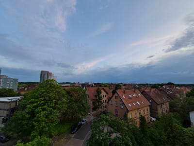This is an aerial view of a residential neighborhood in Brunswick, Germany, likely taken during twilight. The sky is a soft blue with scattered clouds, tinged with pink and purple hues from the setting sun.
The scene is dominated by red-tiled roofs of various houses, interspersed with lush green trees. Some of the houses appear to be older brick buildings, while others are more modern with stucco exteriors. Several cars are visible on the street, including a black sedan and a silver hatchback.
In the background, taller buildings stand out, including a multi-story apartment complex and what appears to be an industrial structure. The overall atmosphere is peaceful and quiet, suggesting a residential area at the end of the day. The presence of trees and residential buildings clearly indicates an urban or suburban setting. There is no visible text in the image.
