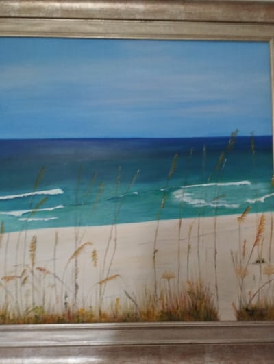 The image is a painting of a beach scene. The sky is a clear blue, transitioning to a darker blue at the horizon where it meets the ocean. The ocean itself is a vibrant turquoise with white waves breaking on a sandy shore. In the foreground, tall, dry grasses are depicted, with some smaller green and yellow plants at their base. The painting is framed by a silver-toned border. There are no people or animals visible in the painting. The painting does not depict any specific location in the city of Zikhron Yaakov, Israel, or any recognizable Israeli landmarks.