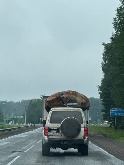 The image shows the rear of a silver Mitsubishi Pajero SUV driving on a wet asphalt road. The vehicle has a large, brown, tarp-covered object secured to its roof rack, possibly camping gear. The license plate reads "X034ME 138 RUS". In the background, to the left, is a green structure resembling a gas station with a white canopy. To the right, a blue road sign points left with Russian text that appears to say "МАРЬЕВКА" (Mar'evka) and "К. ГОРНО" (K. Gorno), indicating Mar'evka as a destination. The environment appears to be rural or semi-rural, with dense trees lining the right side of the road. The sky is overcast and the weather appears to be rainy or recently rained. The overall scene depicts travel on a highway in Russia under cloudy conditions.