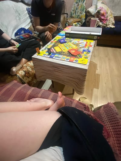The image depicts an indoor scene in Vyatskiye Polyany, Russia, with artificial lighting suggesting it's evening. Several people are gathered, playing a Russian version of the board game Monopoly, titled "МОНОПОЛИСТ РОССИИ" (Monopolist Russia), visible on the colorful game board.

In the foreground, the legs and feet of a person are prominent, resting on a patterned purple and red blanket. This individual appears to be dressed in dark clothing. Around a low table or surface where the game is set up, two other people are seen participating. One person on the left wears a black t-shirt with a white logo, possibly "adidas," and their hands are near the board. Another player, visible on the bottom-right, also in dark attire, holds game pieces or cards.

The game board displays various property squares with Russian text, including "СТАРТ" (START), and numbers like "200," "100," and "60." An open black and white game box is situated next to the board. Other objects include a pink checkered mug and patterned cushions or bedding in the background. The floor is light wood. The scene conveys a casual, social gathering focused on the board game.