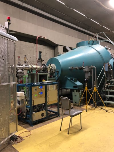 This image captures an indoor scene within what appears to be a scientific laboratory or industrial facility in Agia Paraskevi, Greece. The central subject is a massive, teal-colored industrial apparatus, likely a vacuum chamber, particle accelerator component, or a large experimental vessel, distinguished by its partially conical shape and numerous attached pipes, flanges, and gauges.
To the right of the main apparatus, a metal rack holds several electronic control units featuring illuminated blue digital displays and various buttons, suggesting active monitoring or control. A complex web of wires and cables connects these units to the large vessel and other smaller components. On the yellow floor in the mid-ground, a simple grey metal chair stands, indicating a workstation or resting spot.
Further details include yellow metal support structures, possibly holding a black spherical object, situated near the large apparatus. A red indicator light or object is visible in the bottom-left foreground, close to what looks like a metal grate or cage. The facility features grey walls and a distinctive yellow floor. No individuals are present in the image, and no specific activity is visibly taking place, though the setup clearly suggests ongoing research or industrial operations. The lighting is artificial, typical of an indoor laboratory setting. No discernible text is legible on the equipment.