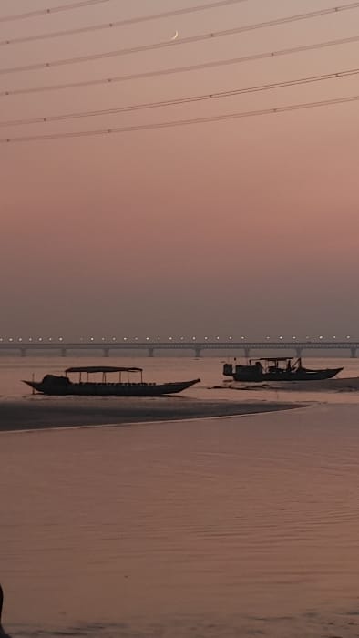 This vertical photograph depicts a twilight scene over a river in Kumarvog, Bangladesh. The upper approximately 60% of the frame shows a sky gradient transitioning from muted orange and pink near the horizon to a faint purple at the top. A slender, white crescent moon is visible in the upper center-left quadrant. Six parallel dark power lines traverse horizontally across the top third of the sky. In the mid-distance, a long, multi-span bridge is illuminated by a series of uniformly spaced bright white lights along its entire length. The lower approximately 40% of the frame consists of calm, reddish-brown water reflecting the sky. Two dark, silhouetted boats are present. The boat on the left is a long, narrow vessel equipped with a canopy, appearing partially beached on a shallow bank. The boat on the right, also featuring a canopy, is floating further out in the water and contains two to three indistinct dark human figures beneath its cover. The immediate foreground displays a dark, indistinct riverbank along the bottom edge.