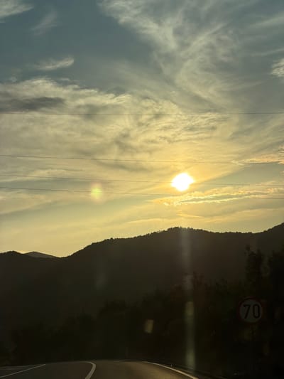 The image shows a landscape in Liješnje, Montenegro, featuring a mountain range silhouetted against a bright, sunlit sky. The sun is partially obscured by clouds, creating a bright glare. Electrical wires stretch across the sky. To the right of the mountain, part of a road and a 70 km/h speed limit sign are visible. The overall scene is bathed in a golden light. The image is vertically oriented.