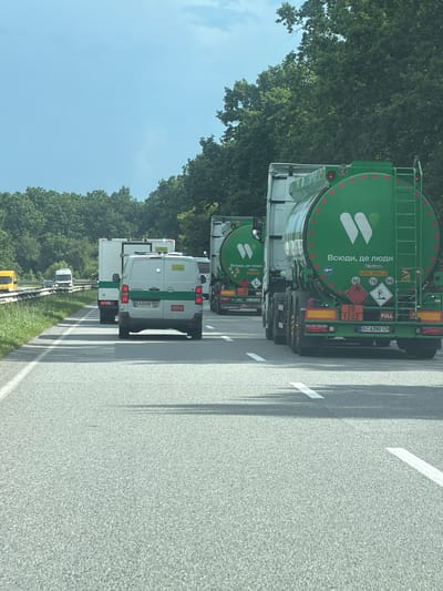 This media file captures a view from inside a vehicle traveling on a highway in Sal'nyk, Ukraine. The main subjects are a convoy of trucks and vans. The most prominent vehicle is a large green tanker truck with the text "Всюди, де люди" (meaning "Everywhere, where people are"). It also displays warning signs for flammable liquids, including "30" and "1202", and "FULL" indicating its load status. Following this tanker are several white vans, one of which has the license plate "KA 0458 TB". Another tanker truck, also green, is partially visible further back, displaying similar text and hazard symbols.

The scene is set on a paved highway with clear lane markings. Trees line the side of the road, suggesting a rural or suburban setting. The sky is a clear, light blue, indicating fair weather and daytime. The angle of the image suggests the vehicles are moving, likely in a procession or as part of a transport operation. The overall impression is one of a functional, perhaps commercial or official, movement of goods or services along a Ukrainian roadway. No people are explicitly visible within the frames of the vehicles.