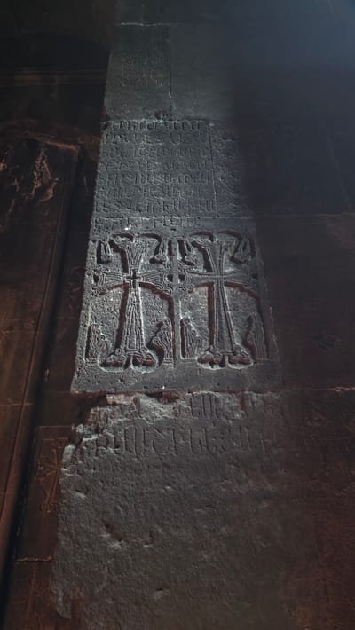 The image shows a stone pillar with carved inscriptions and two elaborate cross designs. The crosses have intricate patterns and are each accompanied by two small, robed figures at their bases. The pillar is dark and appears to be made of rough stone, possibly volcanic rock. The top section of the pillar also features rows of carved inscriptions. The overall lighting suggests an interior setting, possibly a church or ancient structure, with shadows cast across the stone. This is likely a khachkar, a traditional Armenian cross-stone, found at Geghard Monastery in Goght, Armenia.