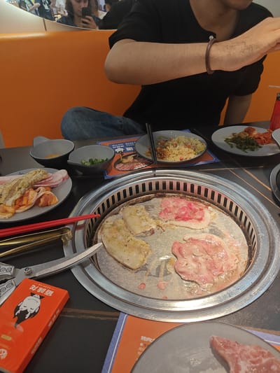 The image shows a close-up view of a table setting at a Korean barbecue restaurant, likely in the Nanhai District of China. The central focus is a round, metal grill where several pieces of raw meat are cooking. Two pieces of meat, appearing to be thinly sliced pork belly, are on the grill, one already showing signs of cooking. Another piece of meat, with more fat marbling, is also on the grill.
Surrounding the grill are various dishes and utensils. To the left, a plate holds more raw meat, some thinly sliced pork and other cuts. Two small bowls are visible: one contains what appears to be seasoning or a dipping sauce, and the other holds chopped green onions. In the background, there are plates of fried rice and what looks like kimchi and seaweed salad.
A pair of red chopsticks and a pair of silver tongs are positioned near the grill, ready for use. To the left of the tongs is an open orange box with a cartoon drawing of a vampire-like character on it and Chinese text. Another blue and white menu or placemat is partially visible in the foreground.
A person, partially visible, is seated at the table, wearing a black t-shirt and blue jeans. Their arm, adorned with a bracelet, is reaching towards the grill. In the reflection of a mirror in the background, a woman is seen taking a picture with her phone. The overall lighting suggests it is daytime, and the setting appears to be indoors, typical of a restaurant. The food and the grill indicate an ongoing Korean barbecue meal. Visible text on the orange box includes "佰味苑".