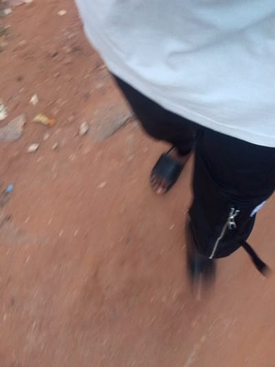The image presents a close-up, downward perspective of a person's lower body, captured in motion. The subject is wearing a plain white t-shirt and black trousers or joggers, which feature a silver zipper detail on a side pocket. One foot is visible, clad in a black slide sandal, with the toes partially exposed.
The setting appears to be an outdoor, unpaved area. The ground is a distinct reddish-brown color, characteristic of laterite soil often found in tropical regions like Orlu, Nigeria. Scattered across the ground are various small debris, including light-colored stones and what might be plant matter or litter.
The entire image exhibits significant motion blur, particularly noticeable on the ground and around the person's feet, strongly suggesting that the person was walking or moving at the time the photograph was taken, or the camera was in motion. The lighting is diffused, indicating daytime, but the precise time of day or specific weather conditions are not discernible. There is no visible text in the image.
