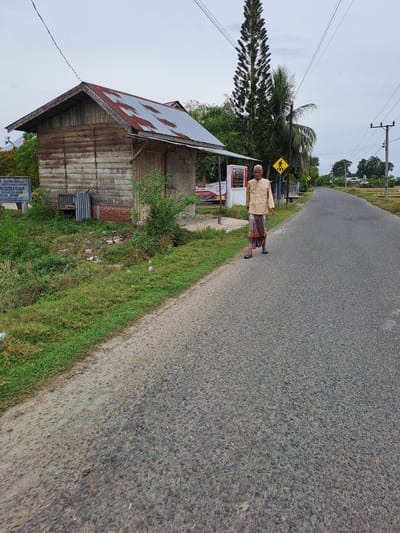 The image depicts an outdoor scene on a paved road in Lancoklancok, Indonesia. A lone elderly man, wearing a light-colored long-sleeved shirt and a patterned sarong, is walking on the right side of the road. He is facing towards the viewer with his head slightly tilted and appears to be holding something in his right hand near his chest.

To the left of the road, there is a small, weathered wooden structure with a rusted metal roof. Adjacent to this building, a blue sign with text and some posters are visible. A small patch of green grass and some scattered debris are present in front of the building.

Behind the man, a tall evergreen tree stands out against the overcast sky. Further down the road, there are other trees, some houses, and what appears to be agricultural land. A yellow pedestrian crossing sign is also visible on the side of the road near the man. The overall atmosphere is neutral, with no discernible interactions between subjects or any indication of specific events.
