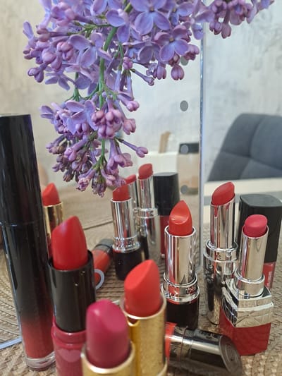 The image shows a still life arrangement of lipsticks and lilac flowers on a reflective surface. The lipsticks are various shades of red and pink, with different packaging designs. The lilac flowers are purple and arranged to the side of the lipsticks. The reflective surface creates multiple reflections of the objects, adding depth to the composition. The background is blurred and indistinct. There is no indication of Bel'kovo, Russia.