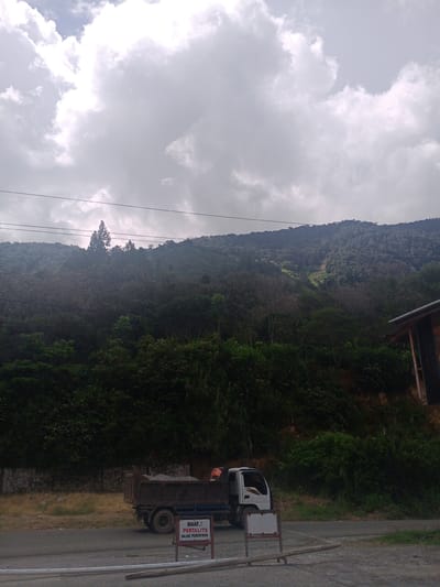 The image shows a scene in the city of Gegarang, Indonesia. In the foreground, there is a dark-colored dump truck parked on the side of a road. The truck is loaded with gravel and appears to be empty of its usual cargo. A person, partially visible in an orange vest, is standing near the back of the truck, possibly overseeing its operation or resting. In front of the truck, a white sign with red text is placed on a stand. The text on the sign reads "MAAF, PERTALITE DALAM PENGIRIMAN." This translates to "SORRY, PERTALITE IS IN TRANSIT."
The background of the image is dominated by a lush, green mountainside covered in dense vegetation. Several power lines are strung across the frame, running horizontally in front of the mountain. The sky above is overcast with dramatic, grey clouds, suggesting potential for rain or a cloudy day. A portion of a wooden building with a dark roof is visible on the far right edge of the image. The overall impression is of a rural or semi-rural environment, possibly a construction site or a transport hub, located in a mountainous region of Indonesia.