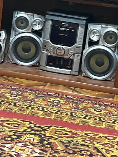 The image displays a multi-component stereo system positioned on a wooden surface, set against a dark background on the left and a vibrantly patterned rug on the right. The stereo system features a silver-grey central unit and two corresponding speakers, one situated above and one below the main unit. Each speaker appears to have at least two drivers: a larger main speaker cone with a yellowish-green tint and a smaller tweeter above it.
The central unit of the stereo system shows a visible disc tray opening on its left side and various controls, buttons, and possibly a cassette deck slot on the right. Faintly visible text on the central unit includes "Panasonic" and "SUPER WOOFER," indicating the brand and a feature.
To the right of the stereo, a section of a traditional rug is visible, characterized by intricate patterns in shades of red, yellow, black, and cream. The rug's design suggests an ornate, possibly oriental or Middle Eastern, style. The wooden surface beneath the stereo appears to be a shelf or part of a furniture unit. The scene is indoors, likely a domestic setting in Jrarbi, Armenia, with no specific activity taking place. The overall impression is that of a functional, older model audio system.