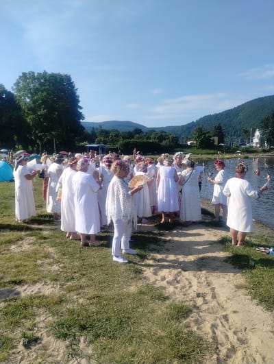 The image depicts a gathering of approximately twenty-five individuals, primarily women, dressed in white attire and adorned with flower crowns, standing on a sandy bank adjacent to a body of water. Some individuals are holding fans. In the background, several people are visible in the water, and there are tents and a stage-like structure further back. The environment includes a body of water, a grassy bank with sand, trees, and rolling hills under a clear blue sky. The scene appears to be taking place in Międzybrodzie Bialskie, Poland, likely during a warm weather event or festival.