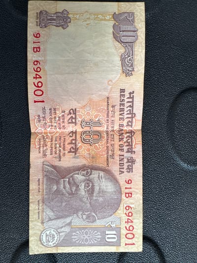 This is an image of an Indian 10 Rupee banknote. The banknote is a light beige color with red and dark brown text and imagery. It features a portrait of Mahatma Gandhi on the left side and the Ashoka Pillar emblem at the top left. The denomination "10" is printed prominently in the top right corner and in the center. The words "RESERVE BANK OF INDIA" are printed vertically in English and Hindi. The banknote appears to be lying on a black, textured surface, possibly a table or mat. There is no indication of the city of Mejaniga, Italy, in this image.
