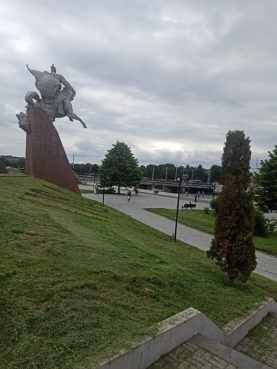 This is a vertical, eye-level, outdoor shot of a monument with a statue in Nazran, Russia. The monument is made of stone and depicts two figures on horseback. The statue is grey and weathered, and the monument base is a reddish-brown stone. In the background, there is a paved walkway with people walking and sitting on benches. There are also trees and lush green grass. The sky is overcast and cloudy, suggesting it is daytime. The overall mood is calm and serene. No visible text is present in the image.