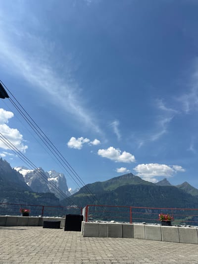 This media file captures a scenic mountain landscape, likely taken from a viewpoint or balcony, with the Swiss Alps as the backdrop. The sky is a vibrant blue, dotted with scattered white clouds, suggesting a clear day. In the foreground, a paved area with stone tiles leads to a red railing. Black seating units are visible behind the railing, and a planter with pink flowers adds a touch of color.

The dominant subjects are the majestic mountains, characterized by their lush green slopes in the mid-ground and snow-capped peaks in the distance. The presence of cable car wires diagonally traversing the frame suggests that this location is either near a cable car station or along a cable car route, a common feature in alpine regions. The time of day appears to be daytime, with bright sunlight casting shadows and illuminating the landscape. The overall mood is serene and awe-inspiring, typical of a breathtaking mountain vista. No visible text is present in the image. The location context of Hasliberg Reuti, Switzerland, aligns with the visual cues of alpine scenery and potential cable car infrastructure.