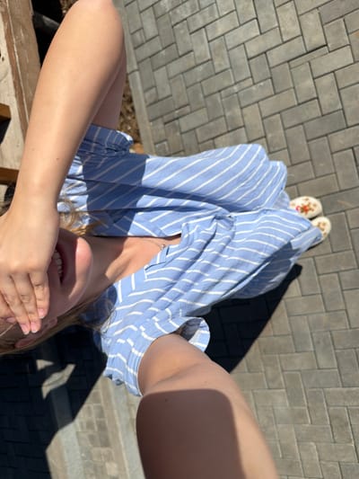 A woman is taking a selfie outdoors in what appears to be Kamennoye, Russia. The image is taken from a high angle, looking down at the woman who is covering part of her face with her hand. She is wearing a blue and white striped dress. Her feet are visible, clad in white slippers. The background consists of a paved area with interlocking gray bricks laid in a herringbone pattern, and shadows are cast by a nearby object.