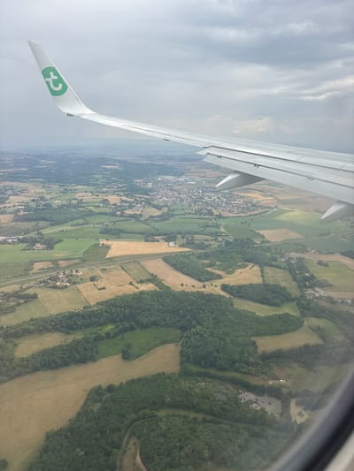 An aerial view from an aircraft depicts the rural landscape surrounding Bonnefamille, France, under an overcast sky. The white wing of a Transavia aircraft, featuring a green 't' logo on its wingtip, extends from the upper left towards the mid-right. Below, the terrain is a mosaic of cultivated fields, presenting in shades of green and yellow-brown, interspersed with dense, dark green forest patches. The commune of Bonnefamille is centrally visible in the mid-distance, identifiable by its cluster of residential and commercial structures with predominantly light-colored roofs. A prominent linear light-colored feature, consistent with a highway (likely the A43 autoroute), traverses the landscape in the mid-ground. Several small, irregular-shaped ponds are visible within forested areas in the foreground. The sky above is uniformly covered by a thick layer of grey clouds. No individuals or specific actions are discernible within the frame.