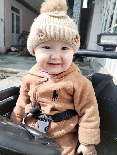 A young infant is shown smiling, seated in a black child's ride-on vehicle or seat, secured by a black safety harness with a plastic buckle. The infant wears a tan knitted beanie with a large pom-pom, three dark brown paw print appliques, and a light brown animal face applique. Underneath, a tan fleece zip-up jacket with the dark brown Ralph Lauren polo player logo on the left chest is visible, with a striped pink and white garment beneath the collar. The background is an outdoor residential setting with a light-colored ground surface. To the left, a light-colored building features a white door and steps with a dark railing. To the right, another light-colored building exhibits multiple white-framed windows with grates. A black rectangular device, possibly a light or speaker, is mounted on a black bar to the right of the infant's head. The scene is located in Limbur, Uzbekistan.