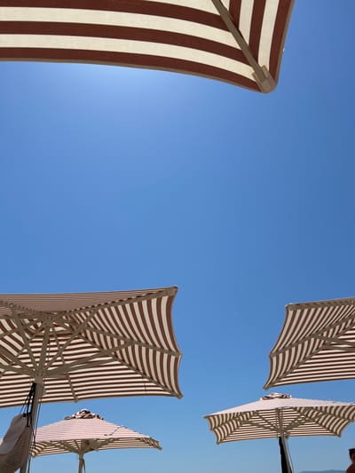 In Troulos, Greece, under a clear blue sky, several striped beach umbrellas are angled upwards. One umbrella is partially visible at the top of the frame, with brown and white stripes. In the lower half of the image, two more umbrellas are open, their striped canopies and supporting structures visible against the sky. To the left, another umbrella is seen from below, with a woven bag and a white hat hanging from its pole. A fourth umbrella is positioned in the lower right, partially obscured by other elements. The scene is set outdoors, suggesting a beach or a similar recreational area on a sunny day.