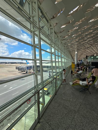 This image depicts the interior of an airport terminal in Bao'an District, China, with a large glass window overlooking the tarmac. Through the window, an airplane is visible parked at a gate, connected to a jet bridge. The terminal interior features a modern architectural design with a high ceiling and extensive use of glass and white structural elements. A row of lounge chairs is arranged along the window, occupied by several people, some of whom are seated and looking at their phones. A young girl stands near the window, looking out. The floor is a speckled grey tile, and the overall atmosphere appears to be one of waiting or transit. The lighting suggests it is daytime.