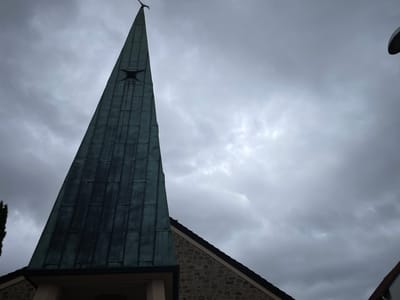 The image shows a close-up, low-angle view of the steeple and roof of a building, likely a church, under a cloudy sky. The steeple is tall and pointed, covered in dark, weathered green metal panels arranged vertically. Near the top of the steeple, a small square window is visible. At the very apex of the steeple, a weather vane shaped like a bird is perched. The roof visible to the right of the steeple is made of stone and tile. The sky is overcast with heavy, gray clouds, suggesting it might be a gloomy day. A lamppost or similar fixture is partially visible in the upper right corner. The scene appears to be in the city of Schorbach, Germany.