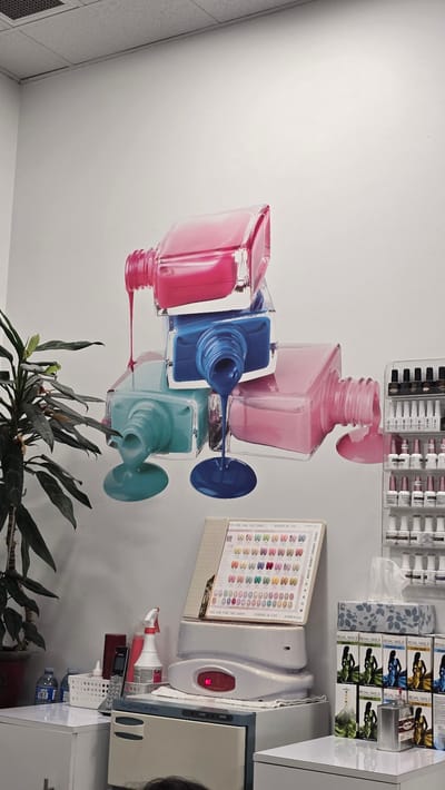 The image depicts the interior of a nail salon in Milton, Canada. A large mural of stacked nail polish bottles, with polish dripping down the wall, is the dominant visual element. In the foreground, a white counter displays various items including a potted plant, a spray bottle, a telephone, a nail polish color swatch display, a nail dryer, a towel warmer, and stacks of nail polish boxes. A shelf unit filled with bottles of nail polish is visible on the right side of the frame. The environment appears to be a clean and organized commercial space. There are no people prominently visible in the scene.