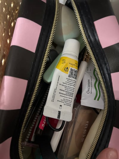 The image shows a close-up of an open black and pink striped makeup bag with a gold zipper. Inside the bag are various cosmetic items. A white tube with yellow and black text is prominently visible, with a QR code and "Medela AG" printed on it. Next to it is a small white packet with green wavy lines and text in a Cyrillic script, which appears to read "Оксолин". Further back, other makeup and toiletries are partially visible. The background suggests the bag is resting on a woven surface. The scene is indoors and lacks any discernible urban or environmental context related to Qoyankus, Kazakhstan.