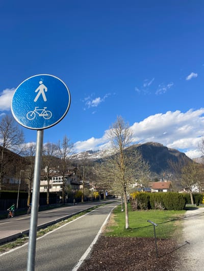 The image captures a shared pathway for pedestrians and cyclists near Sterzing-Vipiteno, Italy. A blue circular sign with white pictograms indicating the shared path is prominently featured in the foreground. The background reveals a landscape with a paved pathway, bordering a grassy area. Trees line the path, some bare and others budding. Houses are visible beyond the trees, and a mountain range with patches of snow rises in the distance under a bright blue sky with scattered clouds. Two cyclists are seen further down the pathway. A solar panel is visible on the rooftop of a house.