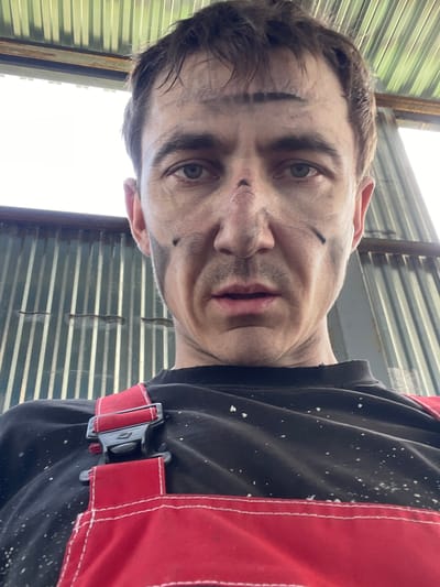 The media file is an image of a man, appearing to be an adult male with fair skin and brown hair. He is wearing a black t-shirt with red overalls over it. The overalls have a black buckle fastening. The man's face is dirty, with smudges of black on his forehead, cheeks, and nose. His expression is neutral. The background consists of corrugated metal walls, suggesting an industrial or workshop setting. There are some white specks on his shirt and the overalls, possibly dust or paint. The lighting suggests it is daytime, as there is a bright light source visible in the upper left of the image, which could be sunlight or artificial lighting. The orientation of the image is rotated. The visible location context is Сокуры, Russia. There is no visible text in the image.