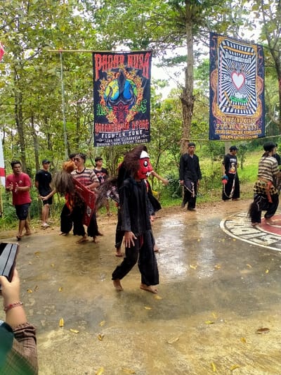 The scene depicts an outdoor cultural or martial arts event taking place on a wet, paved area in Tamansari, Indonesia. A group of approximately 10-12 males are present. In the foreground, a barefoot individual wearing a red-faced mask with white eyes, a white forehead band, a shaggy dark wig, and black long-sleeved attire is in motion. To their left, another barefoot individual in dark pants and a striped shirt with shaggy dark hair moves. On the far right, a barefoot individual in striped pants, a black shirt, and a dark, decorative tail-like attachment is also in motion on a partially visible circular emblem painted on the ground.
In the background, several males observe or participate. Some wear dark, traditional-style clothing, one holding a short staff. Others wear casual attire, including red and black t-shirts and shorts. Two large vertical banners are suspended between trees. The left banner is black, red, yellow, and blue, displaying "PAGAR NUSA," a globe with a trident, and "FIGHTER ZONE." The right banner is black, gold, and white, displaying "PERSAUDARAAN SETIA HATI" with a heart and a stylized emblem. Dense green trees form the backdrop. The ground is wet and light-colored, with scattered yellow leaves, suggesting recent rain. A hand holding a black smartphone is visible in the bottom-left foreground, capturing the event.