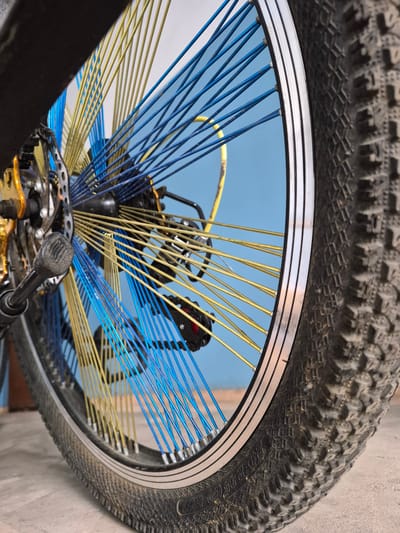 A close-up, low-angle shot captures the rear wheel of a bicycle, emphasizing its colorful spokes. The spokes are a mix of vibrant blue and yellow, creating a striking visual contrast against the muted background. The bicycle's tire is thick and rugged, indicating it's likely meant for off-road use. Partially visible on the left is the bicycle's drivetrain, including the derailleur and chain, with a gold accent on the derailleur. The background consists of a plain blue wall. The scene is indoors, on what appears to be a concrete floor. The image is detailed and sharp, allowing for a clear view of the textures and components of the bicycle. No people are present in the frame. The provided location of Uglegorsk, Russia, does not appear to be directly evident from the visual information within the image itself.
