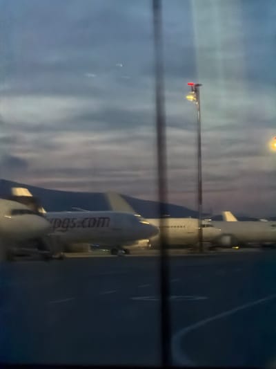 The image displays an airport tarmac at Sabiha Gökçen International Airport (SAW), situated near Tuzla in Istanbul, Turkey, observed during twilight hours. The sky is overcast with tones of blue and grey. Four aircraft are visible on the dark asphalt tarmac. In the central area, a white commercial airliner with "pegasus.com" clearly visible in red text on its fuselage is stationary. To its right, the nose and front fuselage sections of another large, white commercial airliner are visible. Further to the right, the tail section of a third large white airliner is present. In the left foreground, a partial view of a fourth, smaller white aircraft is discernible. Two tall airport light poles equipped with multiple active yellow-white lamps and red top beacons illuminate the scene. A dark mountain range forms a silhouette against the distant horizon. Vertical streaks across the image suggest the view is captured through a window. No individuals or active interactions are visible.