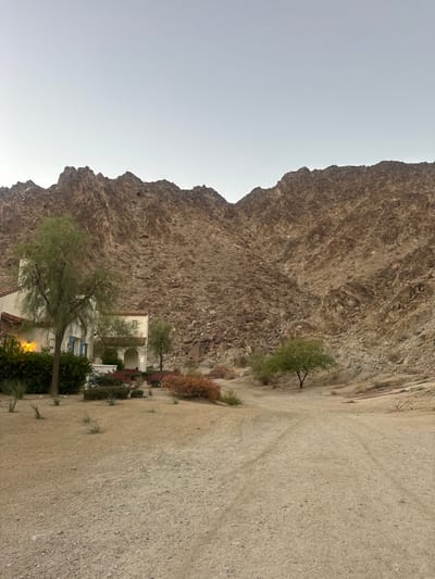 The image displays an arid, mountainous desert landscape at twilight in La Quinta, United States. In the foreground, a light tan, gravelly dirt path with faint tire tracks recedes into the middle ground. To the left, a multi-story residential structure with light stucco walls and a visible section of a reddish-brown tile roof stands. The residence features blue window shutters on its left facade and an arched entryway. Warm yellow light emanates from interior windows. The immediate vicinity of the building is landscaped with various green and reddish-brown desert plants and several small, sparsely leaved trees. The mid-ground and background are dominated by a rugged, rocky mountain range, exhibiting dark brown, tan, and gray geological formations with visible scree slopes. Scattered small trees are present at the base of the mountains. The sky above is clear and light blue-gray. No individuals, animals, or active movements are depicted.