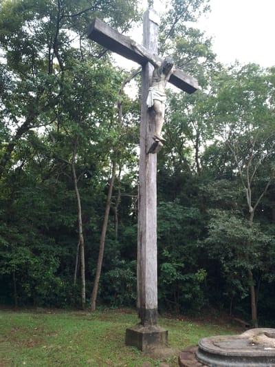 A large, weathered wooden crucifix stands prominently in a natural, wooded setting, likely located in Villarrica, Paraguay. The crucifix features a light-colored statue of Jesus Christ, depicted with a crown of thorns and a white loincloth. The statue appears to be made of a lighter material, contrasting with the dark, aged wood of the cross. The cross itself is tall and stout, fixed into a rectangular concrete base that blends into the grassy ground.

The background is dominated by a dense array of green trees, their foliage creating a lush, verdant canopy that fills most of the upper and middle sections of the image. The trunks of several trees are visible, indicating a mature forest or thick grove. The foreground consists of green grass interspersed with patches of soil or dirt. To the far right, a circular stone structure, possibly a base for another monument or an altar, is partially visible.

The scene appears to be set during daytime, with soft, diffused lighting suggesting an overcast sky or deep shade within the woods. There are no strong, harsh shadows. No specific activity or event is taking place; the image captures a static, serene view of the religious monument amidst nature. There is no visible text in the image. The overall impression is one of solemnity and peaceful contemplation within a natural environment.