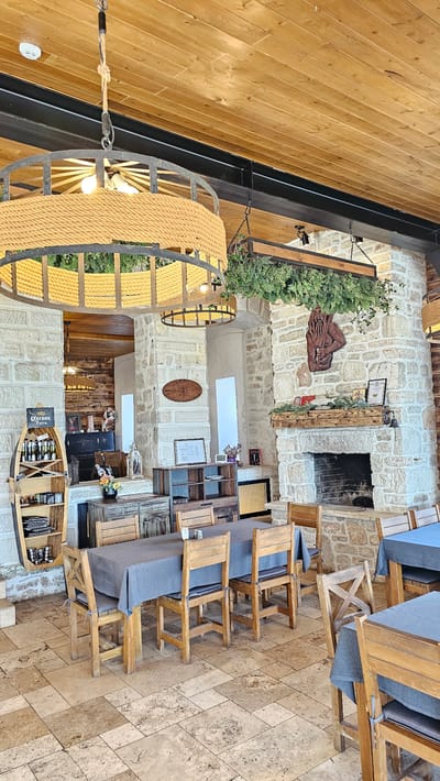 This image shows the interior of a restaurant in Kavarna, Bulgaria. The architectural style incorporates natural stone walls. A large, round chandelier made of rope and metal hangs from the ceiling. Tables and chairs with grey cushions are arranged on a stone-tiled floor. A fireplace is visible on one wall, adorned with decorative items and greenery. Other decorations include wood carvings, potted plants, and boat-shaped shelves filled with wine bottles. The overall ambiance is rustic and traditional.