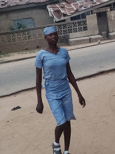 A person with dark skin is walking on a sandy surface adjacent to a paved road in Biliri, Nigeria. They are wearing a blue t-shirt, matching shorts tied at the waist, a blue headwrap, and sandals with white socks. A pair of dark shoes or sandals lie on the ground nearby. In the background, there's a wall with decorative square openings, and buildings with corrugated metal roofs are visible.