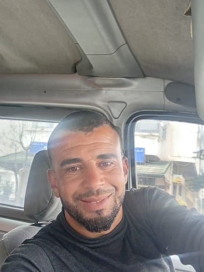 A selfie of a man with a beard in a black shirt taken inside a vehicle in Martil, Morocco, is rotated 90 degrees. Buildings and trees are visible through the vehicle's window. The interior of the vehicle is gray.