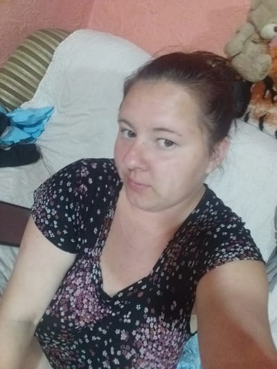 The media file shows a woman lying down in what appears to be a bedroom or living room, given the presence of stuffed animals and bedding. The woman is wearing a black, floral-print top with small white and pink flowers. Her hair is dark and pulled back into a bun. She is looking directly at the camera with a neutral expression. The background consists of a white pillow or cushion and a striped fabric, possibly a couch or bedspread. There are also hints of other soft toys, including a light brown teddy bear, in the upper left corner of the image. The lighting suggests it could be daytime, though indoor lighting conditions are not definitively clear. The overall scene conveys a relaxed, casual atmosphere. The provided location context of Kluhyno-Bashkyrivka, Ukraine, offers geographical information but no specific visual cues within the image directly confirm this location. There is no visible text in the media file.