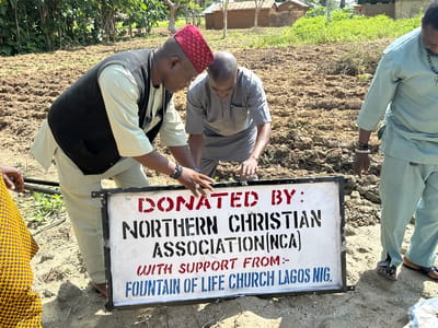 In Afogo, Nigeria, three men are gathered around a white sign with black and red lettering. The sign reads "DONATED BY: NORTHERN CHRISTIAN ASSOCIATION (NCA) WITH SUPPORT FROM:- FOUNTAIN OF LIFE CHURCH LAGOS NIG." One man, wearing a red cap and a dark vest over a light-colored garment, is leaning over the sign, his hands near it. Another man, dressed in a grey outfit, is also leaning over the sign, his hands touching it. A third man, in a light blue garment and wearing bracelets, stands to the right of the sign, looking down. The ground around them is tilled earth. In the background, there are trees and small buildings with thatched roofs. The scene appears to be outdoors, possibly a field or garden.