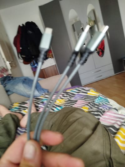 The image depicts an indoor scene, likely a bedroom, located in Ehingen (Donau), Germany. A person's hands are in the foreground, holding several grey USB-C cables. The cables extend upwards towards three distinct white and grey connectors. In the midground, a person is partially visible, lying on a bed covered with a patterned comforter featuring black, white, purple, and yellow stripes. A pair of blue jeans is draped over the bed. Further in the background, a grey wardrobe with silver handles stands against a pale wall. To the left, several jackets and coats are hanging on a wall or hook. The floor appears to be made of wooden planks. The overall lighting suggests artificial indoor illumination.