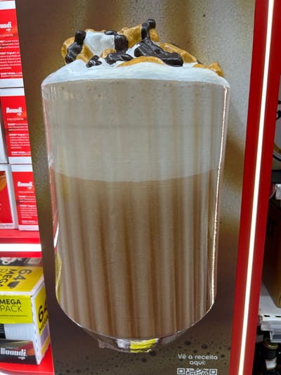 The image shows a close-up of a large, decorative, three-dimensional display of a coffee drink. The drink appears to be a latte or cappuccino with whipped cream, caramel sauce, and chocolate drizzle on top. The display is positioned in front of a wall with a subtle textured pattern. To the left of the display are several red and white boxes, some with the "Buondi" logo and text in Portuguese, suggesting they are coffee products. One box prominently displays "MEGA PACK" in yellow lettering. To the right of the display, a bright red vertical strip with a white illuminated line is visible, possibly part of store shelving or lighting. The lower part of the display features text in Portuguese, "Vê a receita aqui:", which translates to "See the recipe here:", accompanied by a QR code. Based on the presence of "Senhora da Hora, Portugal" in the prompt, it is highly probable that this display is located within a retail establishment in that area, such as a supermarket or a dedicated coffee shop, given the context of coffee product packaging. No people are visible in the image.