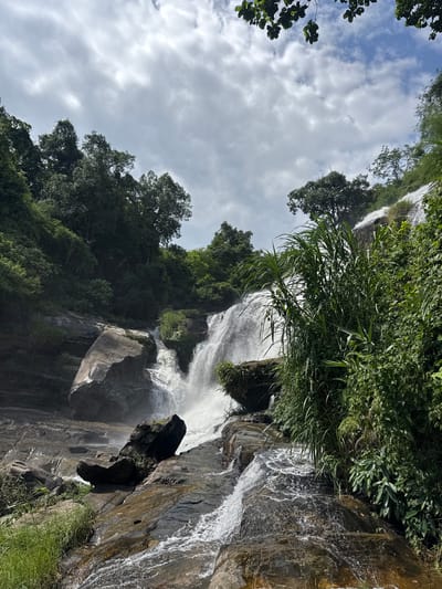 This image depicts a cascading waterfall in Ban Namtok Mae Klang, Thailand. The scene is dominated by lush green vegetation and large rocks, characteristic of a natural jungle environment. The waterfall itself is a prominent feature, with water flowing powerfully over multiple tiers. The surrounding trees are dense and varied, with some reaching high into the sky. The sky above is partly cloudy, with a mix of blue sky and white clouds. The overall impression is one of a serene and wild natural landscape.