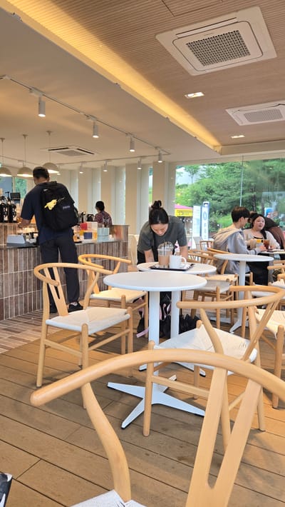 The image captures the interior of a modern cafe, likely located in Gwangmyeong, South Korea, with large windows offering a view of greenery outside. The cafe features light-colored wooden floors and a ceiling with recessed lighting and air conditioning units. Several light-colored wooden chairs with woven seats surround white circular tables.
In the foreground, a wooden chair with a distinctive curved back is prominently featured. In the mid-ground, a woman in a dark-green t-shirt is leaning over a table, seemingly preparing or clearing drinks. On the table are two drinks, one in a clear plastic cup with a straw and another in a white mug.
Behind her, a man with a black backpack adorned with a yellow plush toy is standing at a counter, appearing to be interacting with a coffee machine. Further back, other patrons are visible, including a couple sitting at a table and another person near the counter. The overall atmosphere suggests a relaxed daytime setting in a cafe.