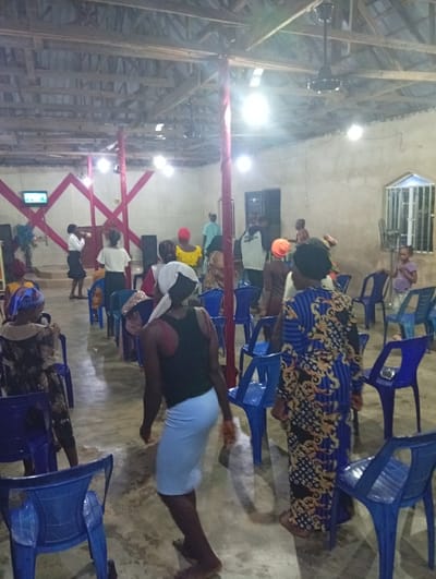 An interior shows a gathering, possibly a church service, with people standing and seated on blue plastic chairs. The room features a high, exposed wooden beam ceiling with bright overhead lights and a ceiling fan. Decorative red fabric adorns a wall, and a small screen is visible. The atmosphere is casual and lively.