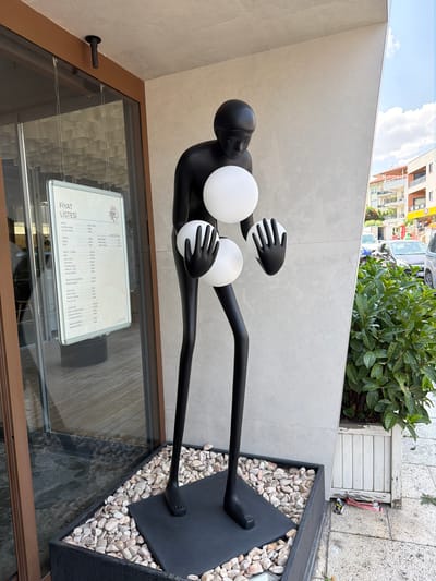 An outdoor shot captures a striking black sculpture of a human-like figure, standing tall and slender with long, thin legs and arms. The figure is depicted holding two large, white spheres, one in each hand. The sculpture is positioned in a planter filled with small, light-colored stones, set against a textured, light-colored wall. To the left of the sculpture, a glass door reveals a glimpse of an interior with wooden flooring and a sign displaying Turkish text, which appears to be a price list. The upper left corner of the image shows a ceiling with a dark fixture. To the right of the sculpture, lush green foliage spills out of a weathered white planter, and beyond that, a street scene with buildings and cars is visible under a partly cloudy sky. The location is identified as Denizli, Turkey, suggesting a commercial or public space.