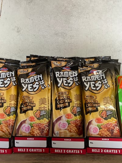 This media file showcases a stack of "Ramen Yes" instant noodle packages, specifically the "Hakata Chicken Ramen" flavor. The packaging is predominantly black and gold, featuring a stylized bowl of ramen with chicken, eggs, and a pink swirl garnish. The brand name "Ramen Yes" is prominently displayed in large, bold letters, with Japanese characters integrated into the design.
The packages are arranged vertically in a stack, suggesting they are being displayed for sale or storage. Each package also has a red strip on the right side with white text that reads "BELI 2 GRATIS 1" (Buy 2 Get 1 Free), indicating a promotional offer. The text "HARGA" (Price) and "Rp." (Rupiah, the Indonesian currency) are also visible, further reinforcing the retail context. Additionally, a "HALAL" certification logo is present on some packages.
The background is a textured, light-colored surface, possibly a countertop or floor, with some subtle grey speckling. The lighting appears to be indoor, casting a slight shadow on the lower parts of the packages. There are no people or distinct environmental clues to suggest a specific time of day or weather. The overall impression is a retail product display, likely in a store or market in Indonesia, given the currency and promotional text.