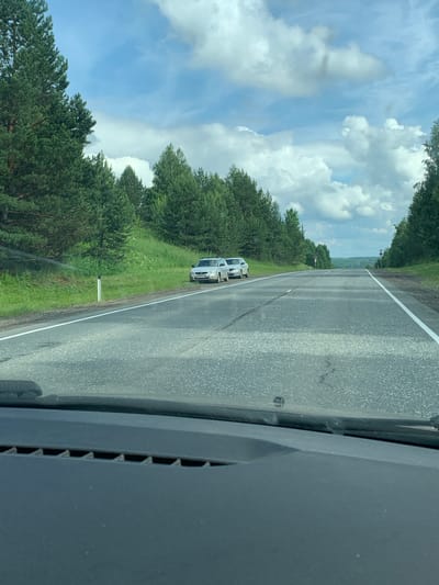 The image shows a view from inside a car looking out at a road in the city of Sudak, Russia. The road is asphalt and has white lane markings. On the right side of the road, there is a grassy embankment with trees. On the left side of the road, there are two silver cars parked on the shoulder. One car is a hatchback, and the other appears to be a station wagon. The sky is blue with white clouds, and the sun is shining. There are trees and hills in the distance. The overall scene suggests a scenic drive on a summer day.