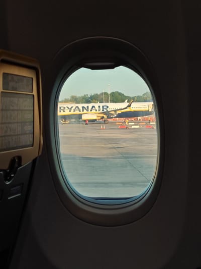 From the perspective of an airplane passenger looking out the window, a Ryanair aircraft is visible on the tarmac. Ground crew are working around the plane. The registration G-RUKE is visible on the aircraft. The location is likely at an airport within Uttlesford, United Kingdom. The interior of the passenger plane with a foldable tray and interior panel is also visible.