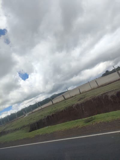 The scene depicts a rural landscape in Kisima ward, Kenya. A paved road is visible in the foreground, with a white dividing line. Adjacent to the road is a grassy embankment that leads to an elevated field. A wire fence runs along the field's edge, supported by wooden posts. The field appears cultivated, potentially agricultural land. In the distance, a treeline marks the horizon, with a cloudy sky overhead. A small patch of blue sky is visible through a break in the clouds. There are no people visible in the frame. The overall impression is one of a rural agricultural area under overcast skies.
